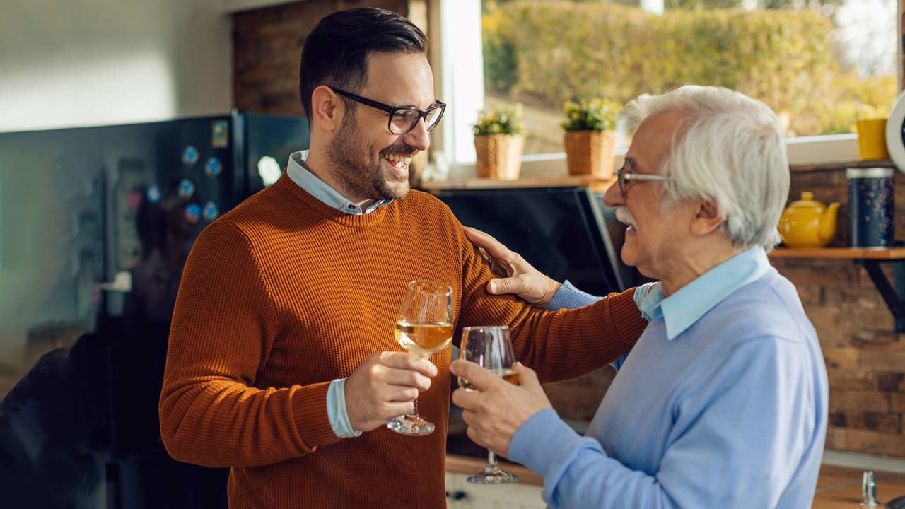Happy man and his mature father toasting with wine talking in the kitchen.