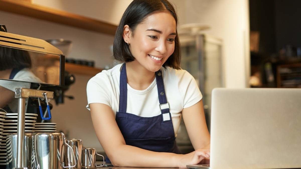 Portrait of smiling asian barista, cafe owner entrepreneur, working on laptop, processing orders on computer, standing behind counter.