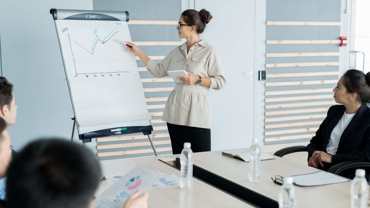 Young sales coach with hair bun wearing glasses holding presentation and explaining graph to business people at training class
