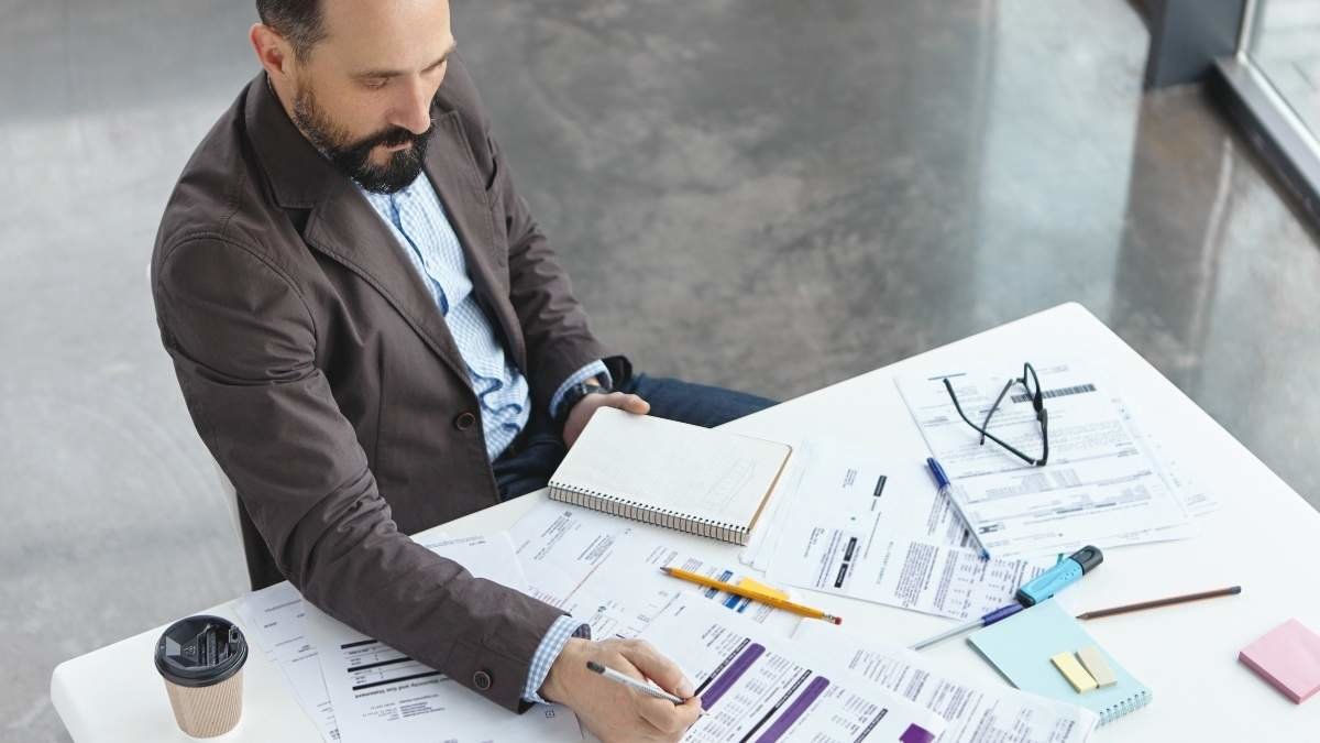 Top view of professional lawyer studies new difficult case, reads attentively papers, makes notes, drinks coffee, involved in working process. Male finacial inspector prepars report, signs documents.