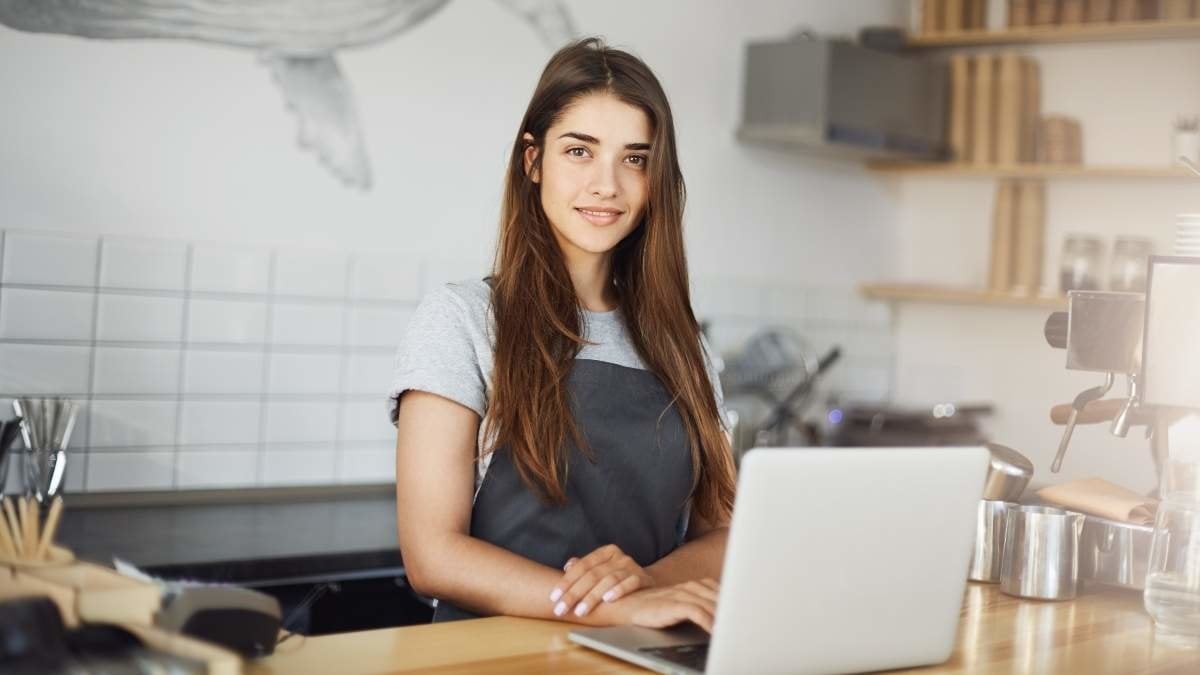 Young female barista using a laptop computer at her work in cafe. Happy employee looking at camera smiling.