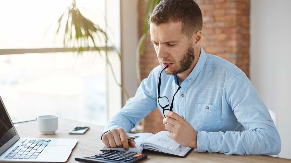 Close up of unshaven serious good-looking finance manager spending morning in office, holding glasses in mouth, looking at calculation results with thoughtful expression