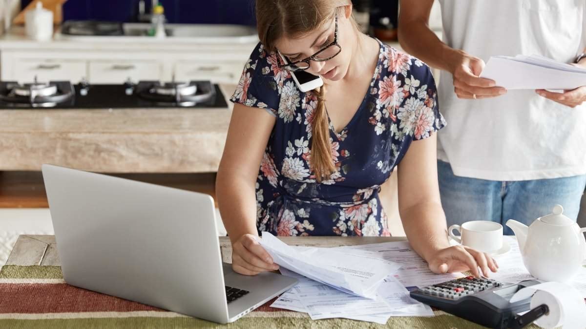 Indoor view of beautiful woman plans family budget, calculates figures and works with documentation, has busy telephone conversation, sits in front of opened laptop at kitchen table, man stands behind