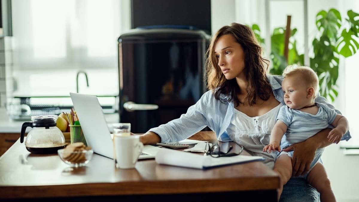 Young working mother using computer while holding her baby and planning home finances.