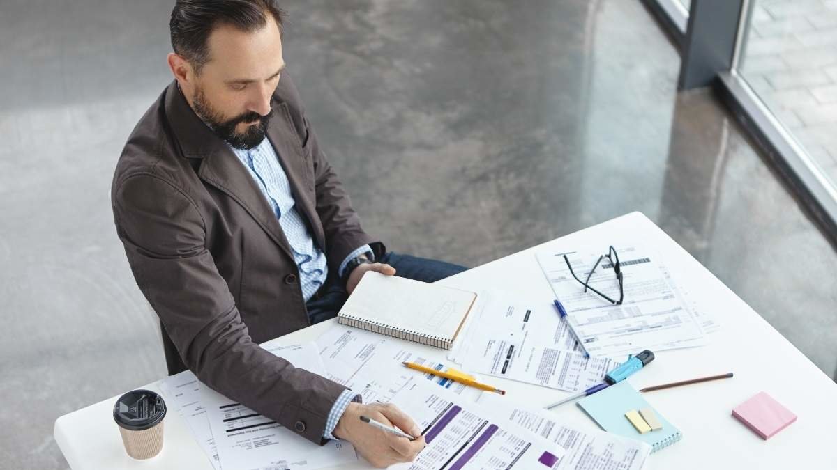 Top view of professional lawyer studies new difficult case, reads attentively papers, makes notes, drinks coffee, involved in working process. Male finacial inspector prepars report, signs documents.