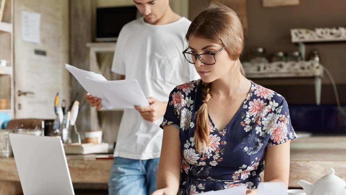 Serious female dressed in blouse, looks attentively at screen of laptop computer, holds documents, count expenses and taxes, prepares financil report or manages family budget together with husband