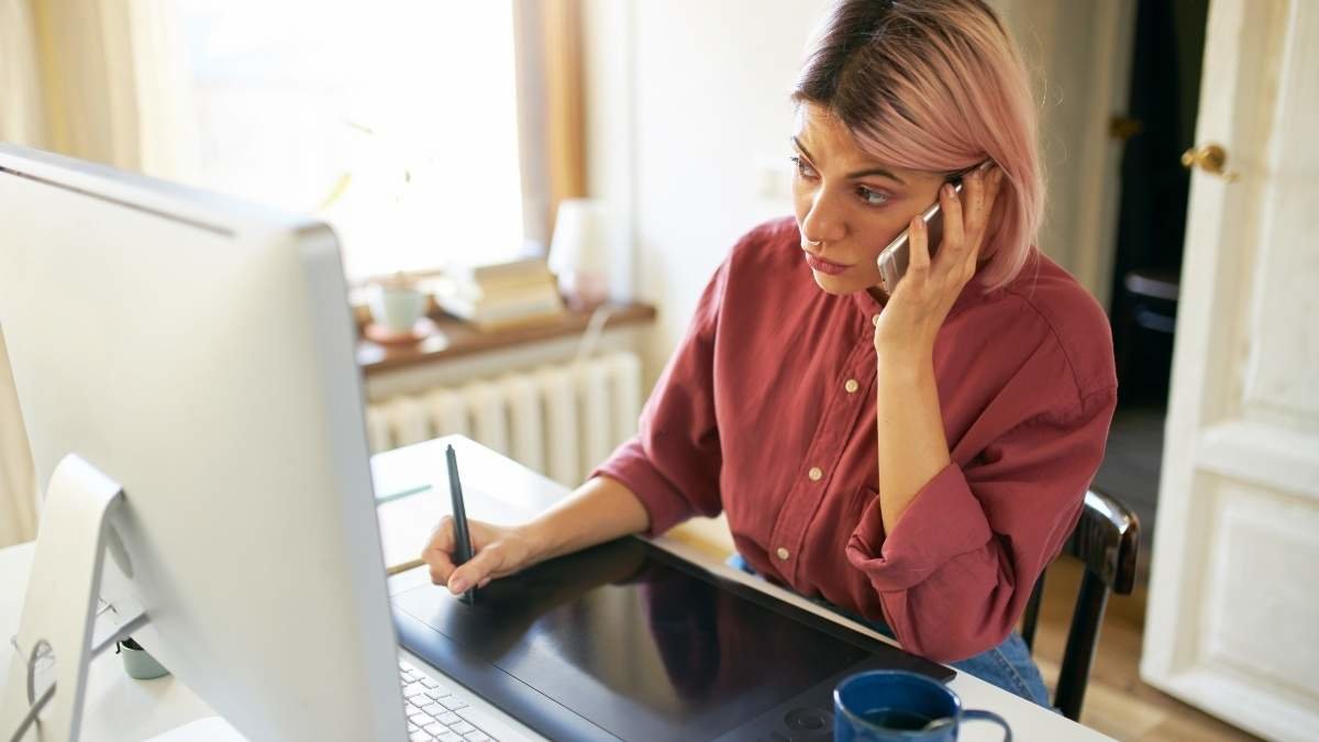 High angle view of serious stylish young woman web designer multitasking while working from home, drawing using graphic tablet, stylus, talking to client on mobile phone. Technology and creative work