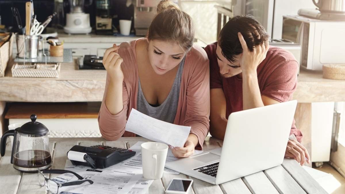 Young couple doing paperwork in kitchen: frustrated woman reading document together with her husband who is holding his head in desperation, sitting at table with laptop, papers and calculator